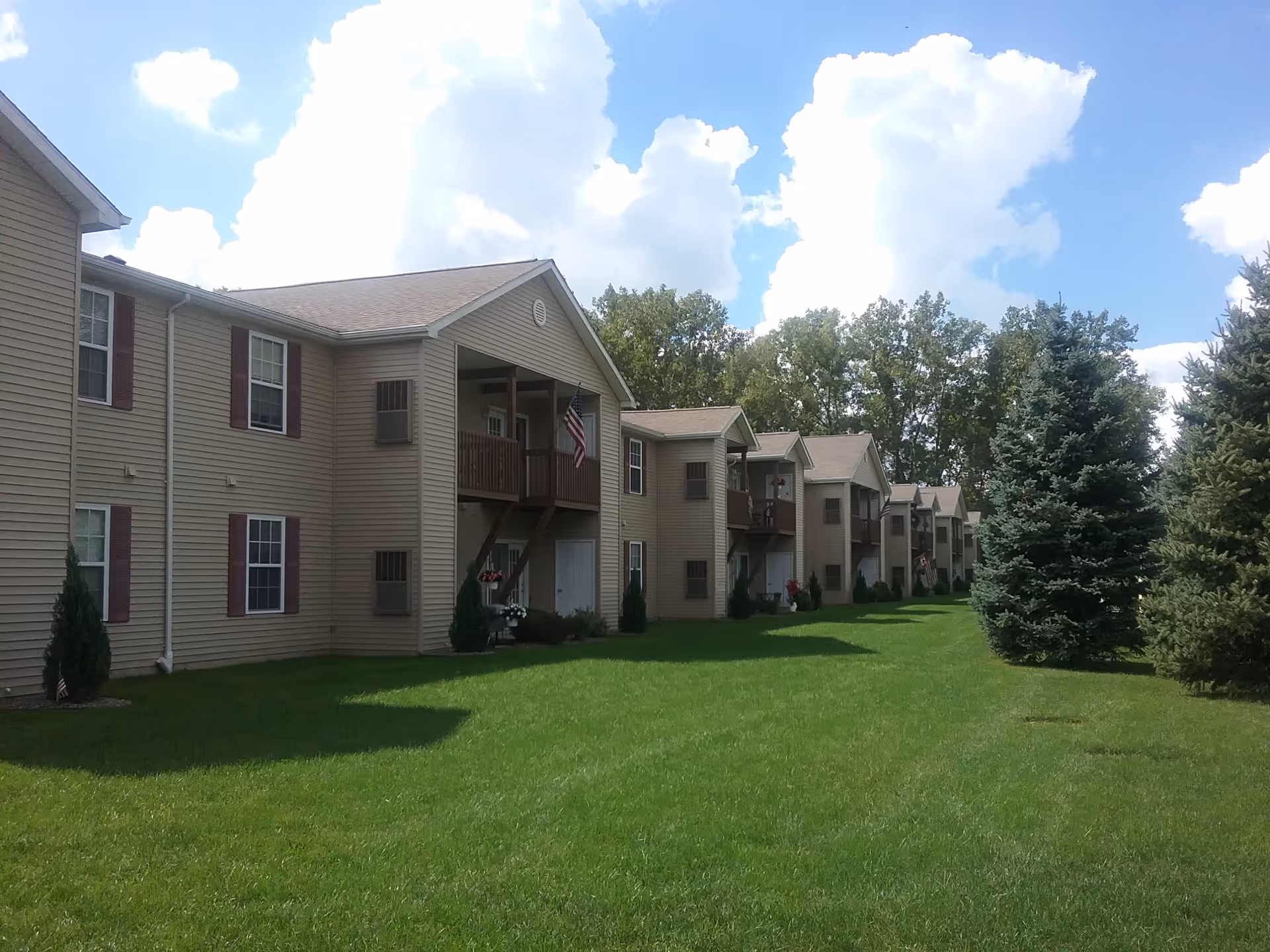 Exterior view of a senior apartment complex with beige siding and brown shutters. The buildings have balconies with American flags hanging. There is a well-maintained green lawn and several tall evergreen trees. The sky is partly cloudy with large white clouds.