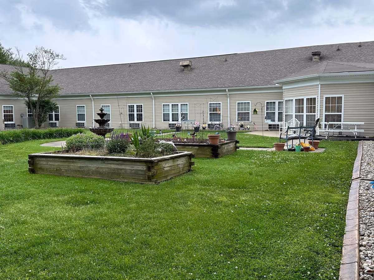 Outdoor garden area at Tender Touch Heart & Home with raised wooden flower beds, a multi-tiered water fountain, green grass, and patio furniture near the building. The building has beige siding and multiple windows with white trim.