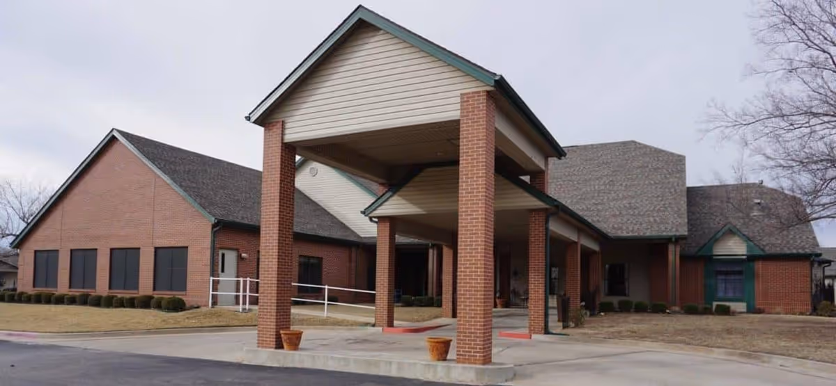 Exterior view of a senior living facility building with a covered entrance supported by brick columns, surrounded by a paved driveway and some landscaping with bushes and grass under an overcast sky.