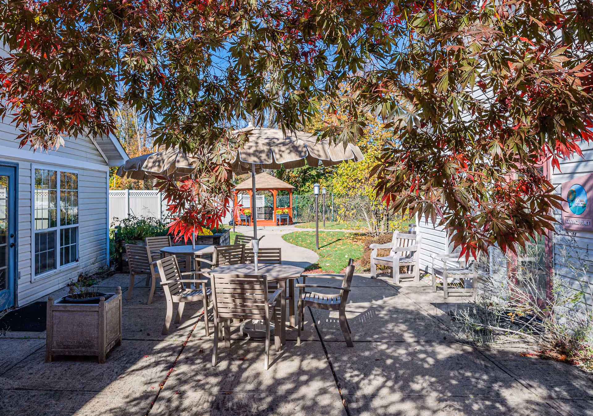 Outdoor patio area with several wooden tables and chairs, one table shaded by a large umbrella. The patio is surrounded by buildings with white siding and blue doors. There is a paved walkway leading to a gazebo in the background, and trees with red and green leaves providing shade.