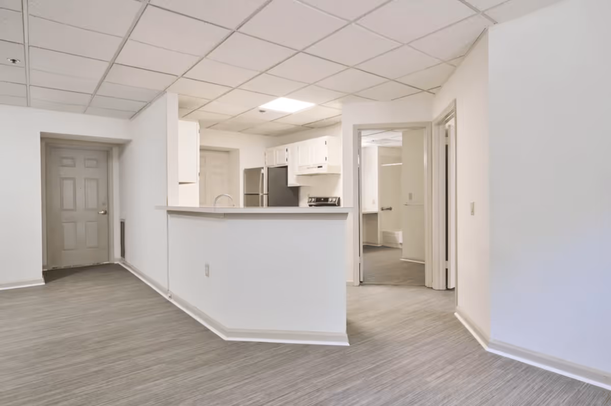 Interior view of a senior living facility showing a small kitchen area with white cabinets, a stainless steel refrigerator, and an electric stove. The kitchen is partially enclosed by a half wall with a countertop. The flooring is a light gray wood-like material, and the walls are painted white. There are doorways leading to other rooms, and the ceiling has a grid pattern with recessed lighting.
