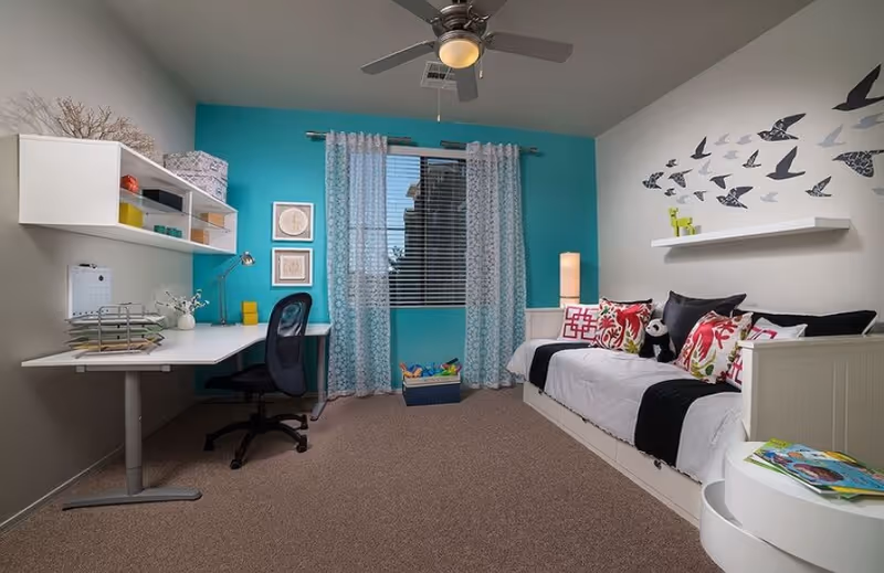 A bright and tidy bedroom with a white daybed adorned with colorful pillows and a stuffed panda. The wall behind the bed is painted turquoise and decorated with a flying bird decal and a white floating shelf holding a small green figurine. Opposite the bed is a white desk with a black office chair, a desk lamp, and organized shelves with various items. A window with patterned curtains lets in natural light, and a ceiling fan with a light fixture is mounted above.
