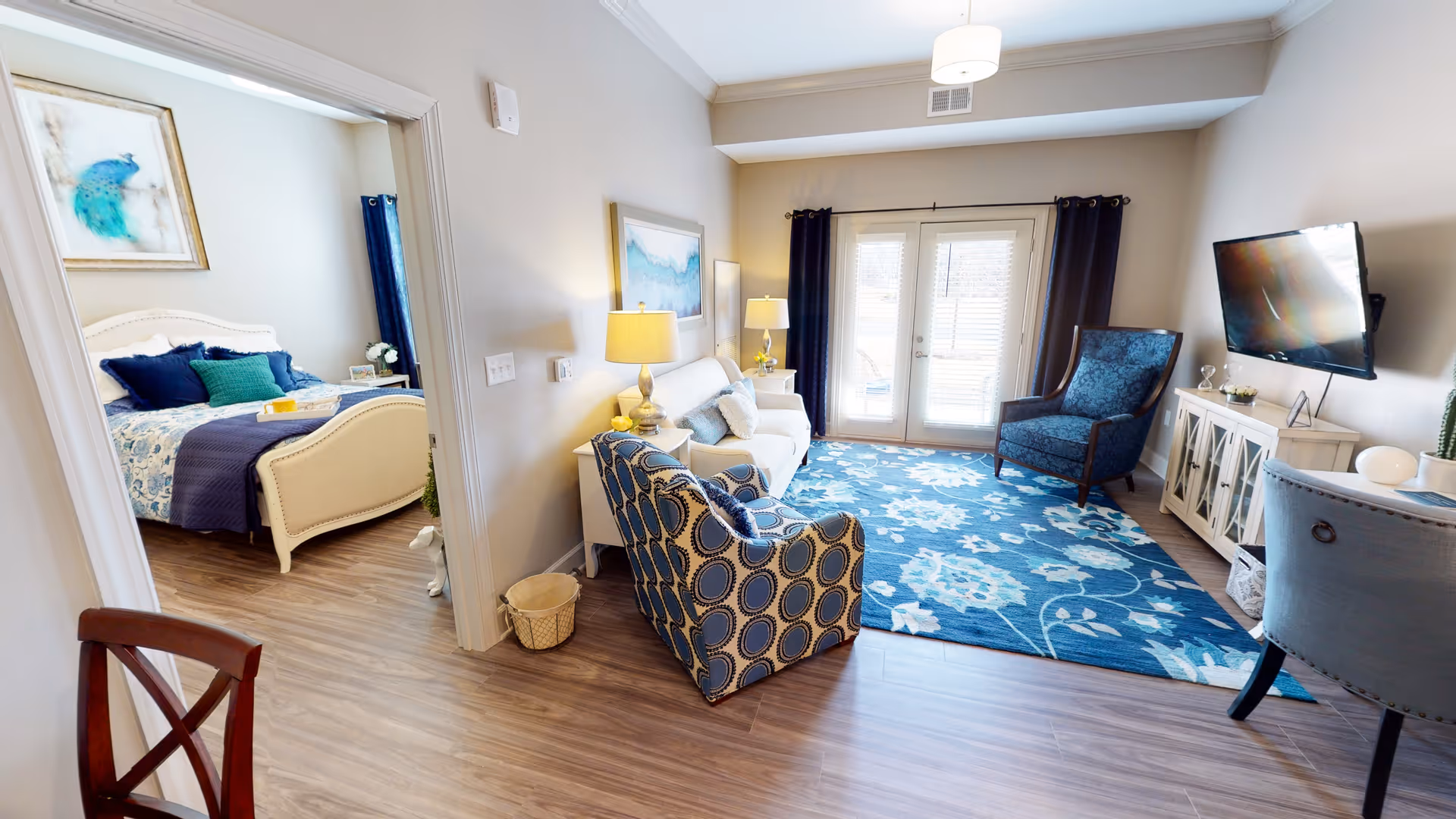 A bright and cozy living room with a blue floral rug, a white sofa, a patterned armchair, a blue armchair, and a wall-mounted TV. The room has double glass doors with dark curtains leading outside. To the left, an open doorway reveals a bedroom with a white bed frame, blue and green bedding, and a framed peacock artwork on the wall.