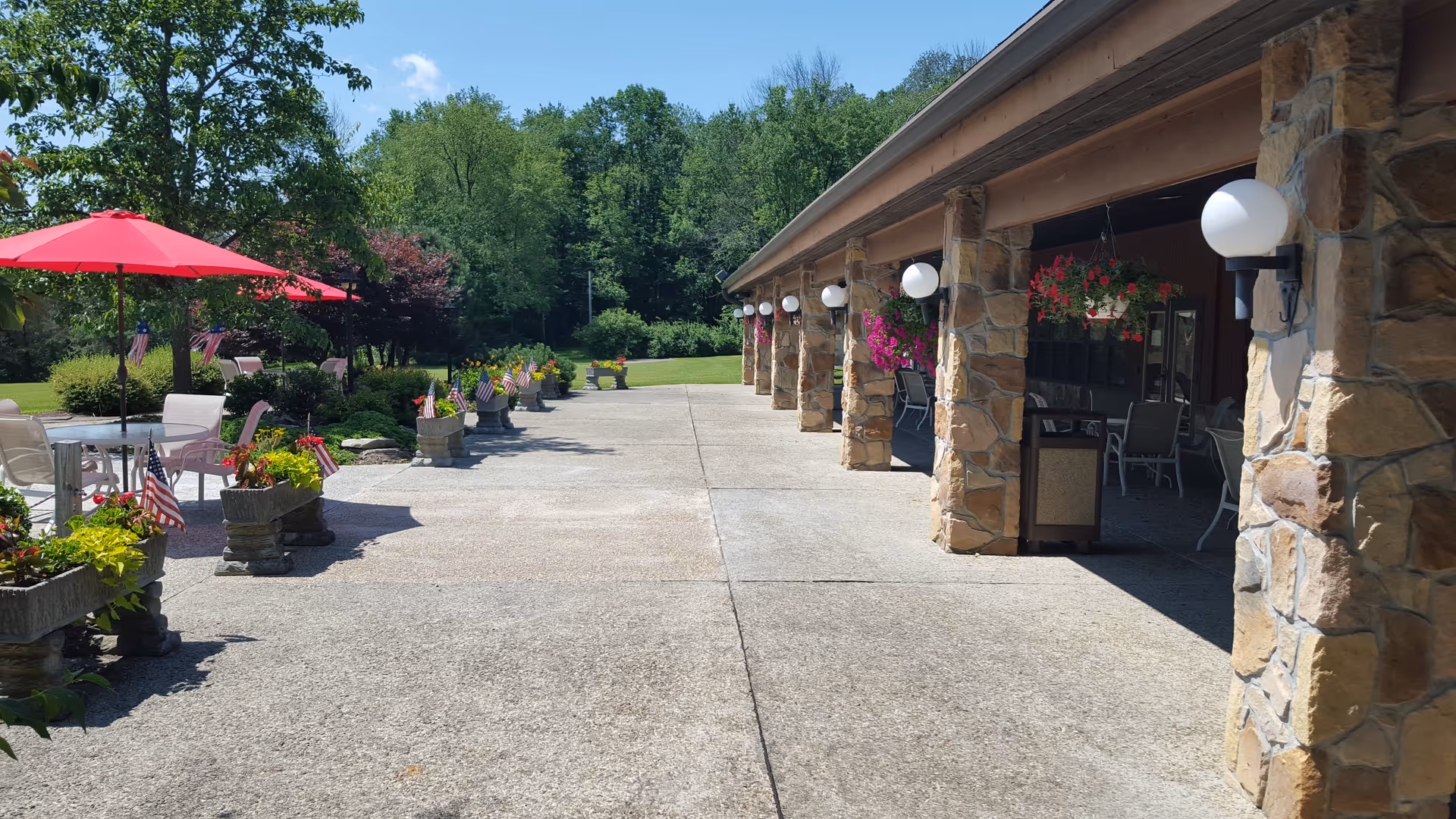 Outdoor patio area with stone pillars and hanging flower baskets on the right side, and several tables with chairs and red umbrellas on the left side. The patio is decorated with flower pots and small American flags, surrounded by green trees and grass under a clear blue sky.