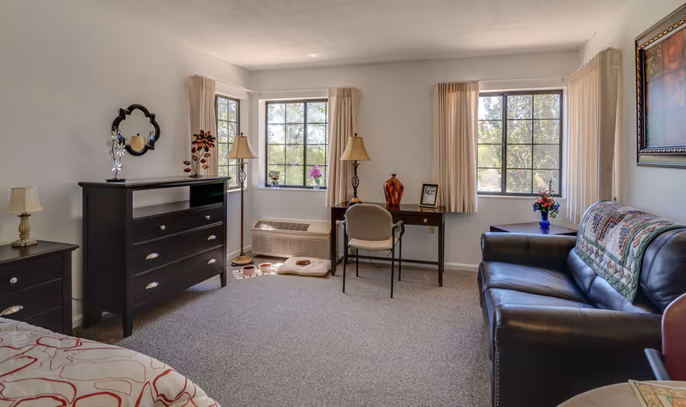 A bright and cozy senior living room with two large windows letting in natural light. The room features a black leather sofa with a quilt draped over the back, a wooden desk with a chair, two table lamps, a dresser with a decorative mirror above it, and a small side table with a lamp. The walls are white, and the carpet is light gray. There are decorative items including a vase with flowers, a framed picture, and a floor lamp with a floral design.