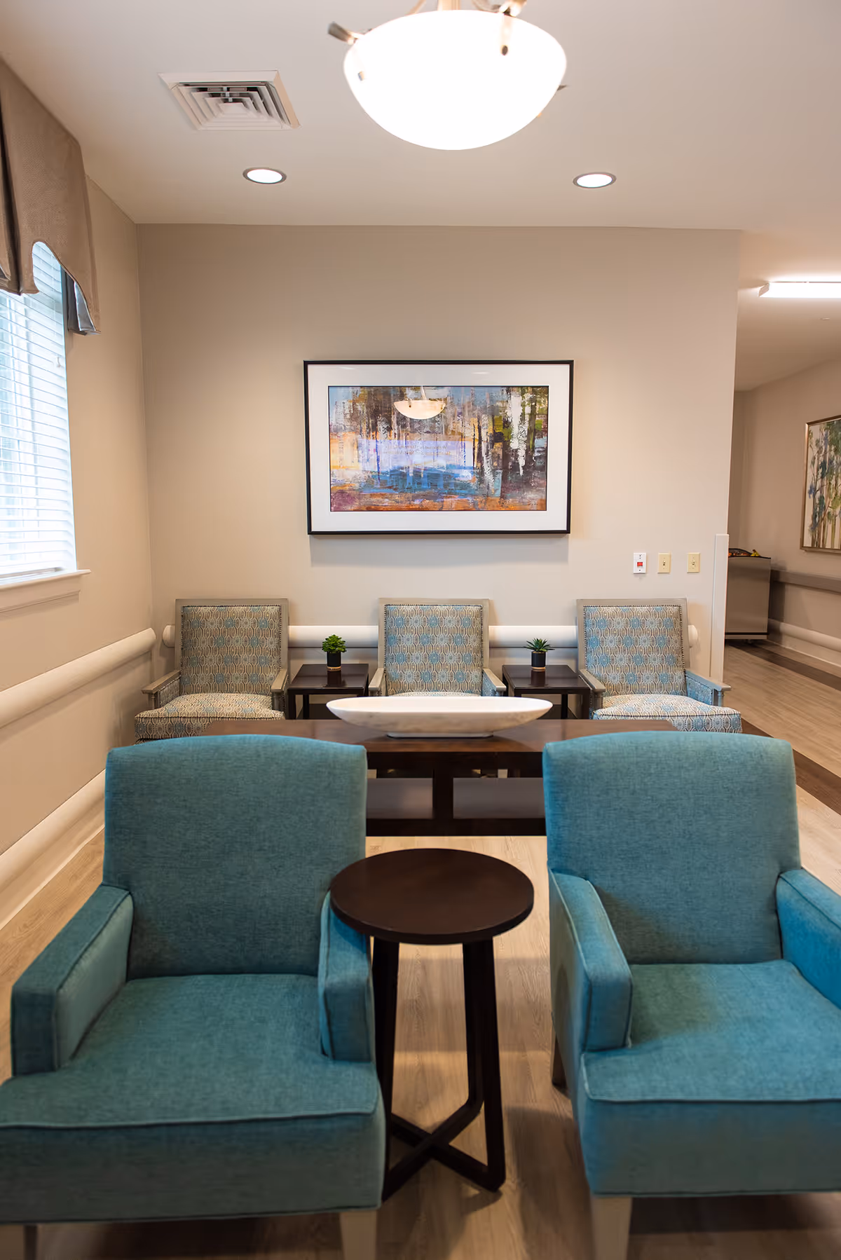 A seating area in a healthcare facility with two teal armchairs in the foreground separated by a small round wooden table. Behind them are three patterned armchairs with small potted plants on side tables between them. A framed abstract painting hangs on the beige wall above the chairs. The room has light wood flooring and a window with blinds on the left side.