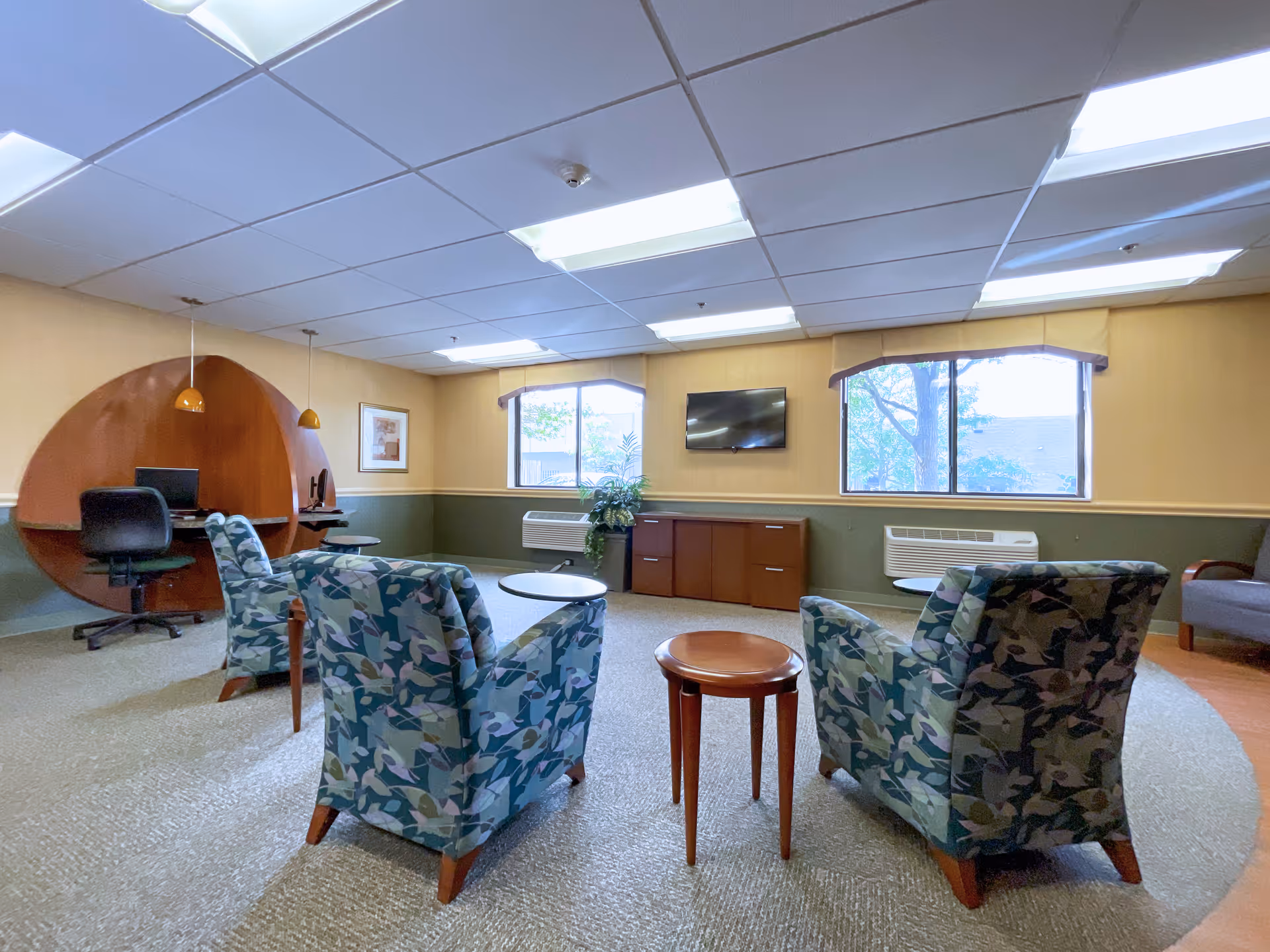 A cozy common area with patterned armchairs arranged around small wooden tables, a wall-mounted TV, two windows with views of trees outside, and a unique wooden workstation with two office chairs and pendant lights.