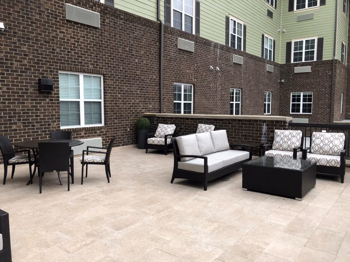 Outdoor patio area with beige tiled flooring, featuring a seating arrangement including a cushioned sofa, two cushioned armchairs with patterned fabric, a glass-top coffee table, and a round table with four chairs. The background shows a brick and green-paneled building with multiple windows.