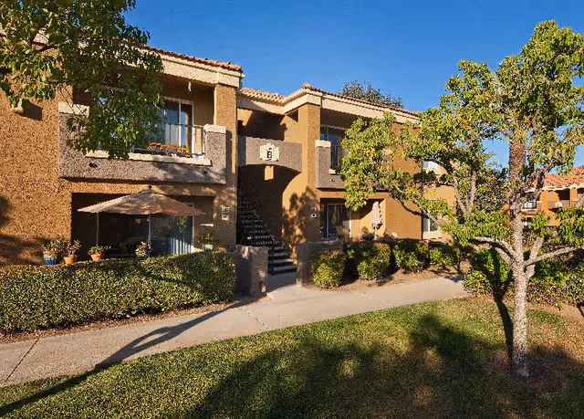 Exterior view of a two-story residential building with balconies and a central staircase labeled with the letter B. The building is surrounded by green bushes, trees, and a well-maintained lawn under a clear blue sky.