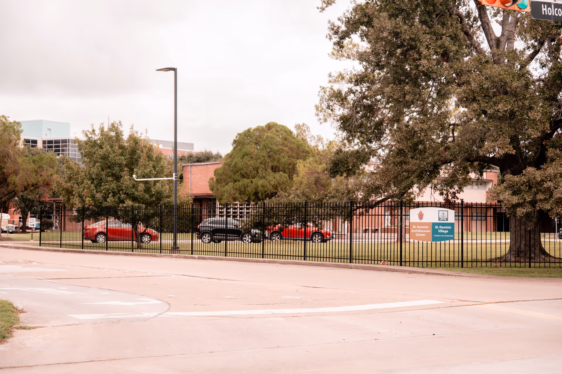 Exterior view of St. Dominic Village facility showing a fenced grassy area with trees, parked cars, and a sign indicating St. Dominic Archdiocesan Center and St. Dominic Village. The scene includes a street intersection with a traffic light and street signs.