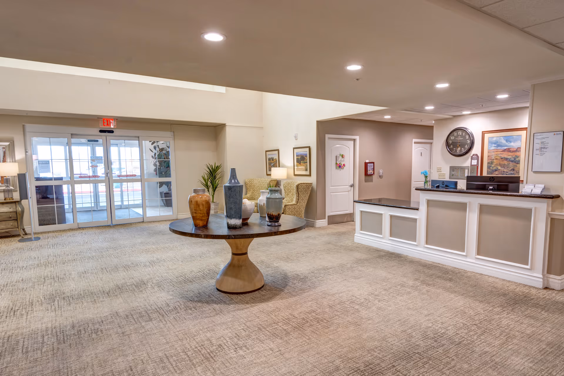 Spacious reception lobby with a round centerpiece table displaying vases, seating area, front desk, and glass entrance doors.