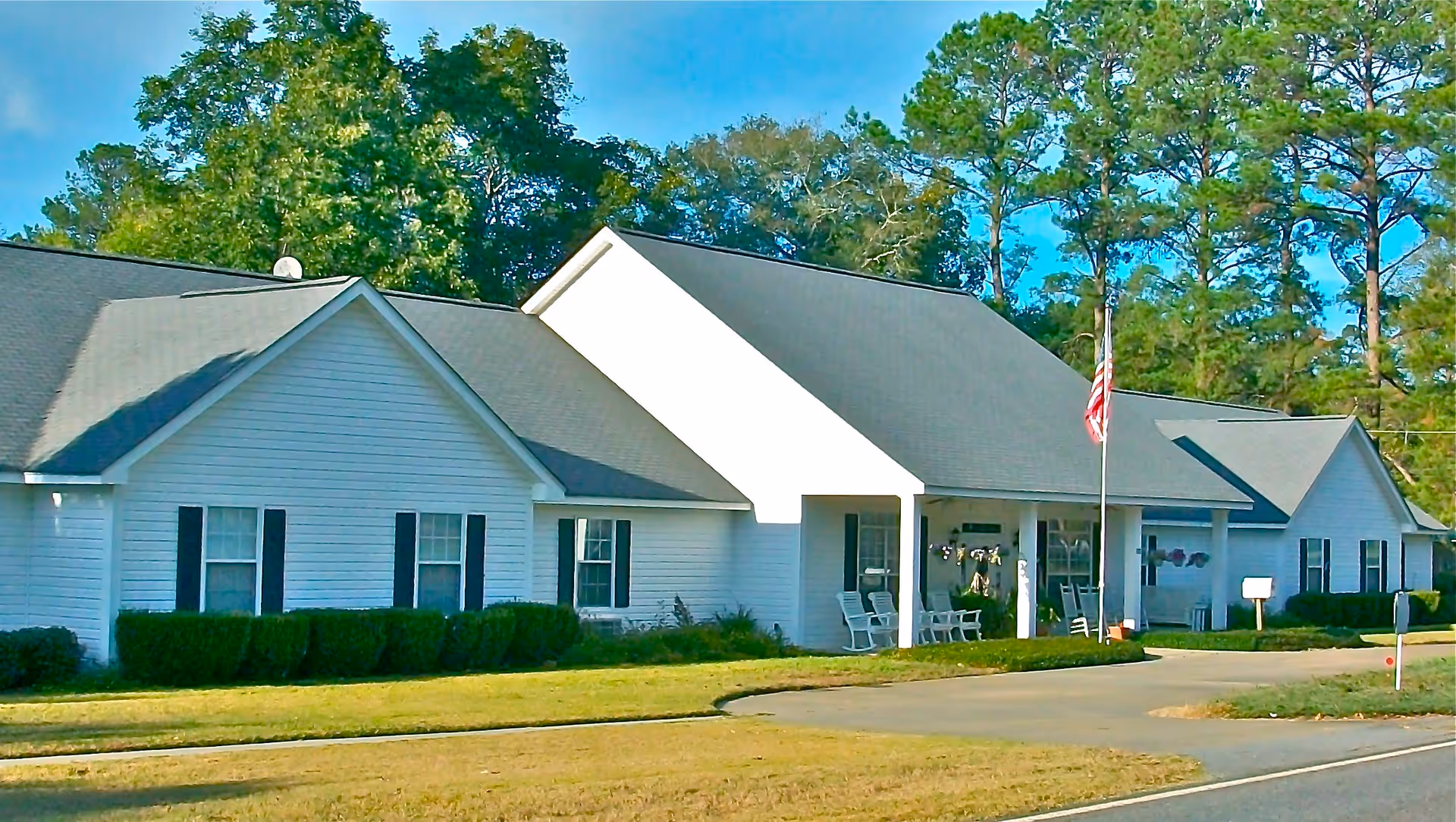 Exterior view of a single-story white building with a dark gray roof, surrounded by green bushes and trees. The building has multiple windows with black shutters and a covered porch area with white rocking chairs. An American flag is displayed on a flagpole near the entrance. The sky is clear and blue.