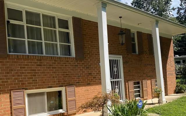 Front of a brick ranch-style house with white porch columns, windows with brown shutters, a decorative metal storm door, potted plants and an ADT security sign on the lawn.