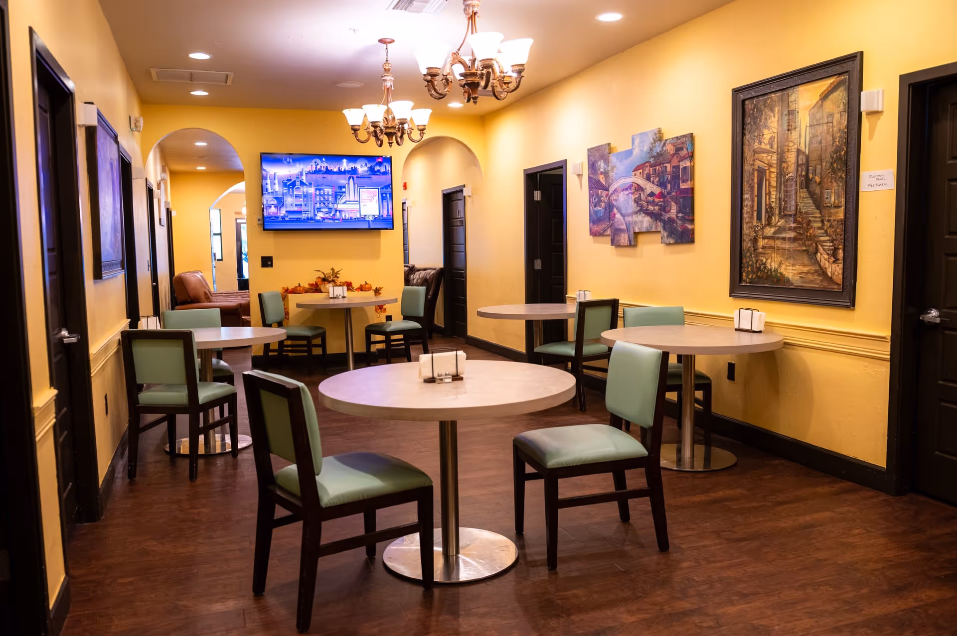 A cozy dining area with round tables and green cushioned chairs set against yellow walls. The room features decorative paintings on the walls, two chandeliers hanging from the ceiling, and a flat-screen TV mounted on the far wall. The floor is wooden, and the space has a warm, inviting atmosphere.