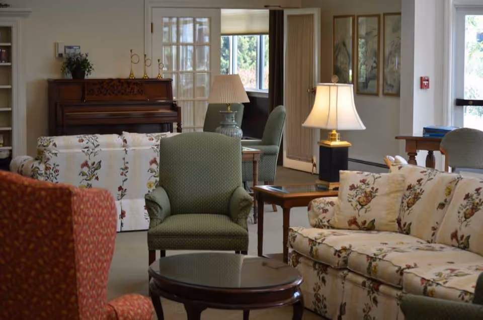 A cozy living room area with floral patterned sofas, green upholstered armchairs, a wooden coffee table, and a table lamp. In the background, there is a piano, a side table with another lamp, framed artwork on the wall, and a window letting in natural light.