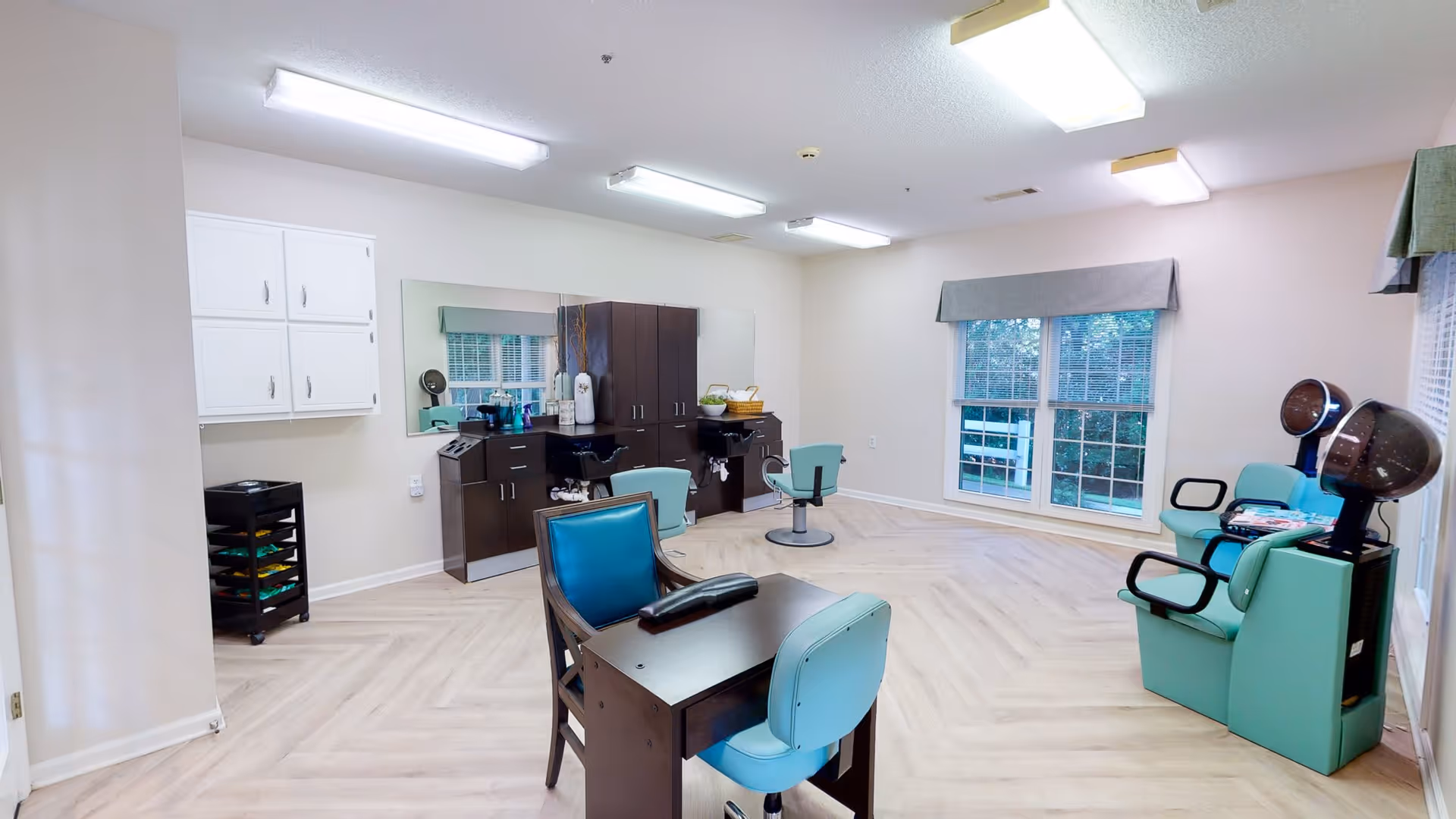 A spacious salon room with light wood flooring and white walls. The room features salon chairs, a manicure table with two chairs, hair drying stations with hooded dryers, and cabinets with a large mirror above them. Large windows with gray valances allow natural light to enter the room.