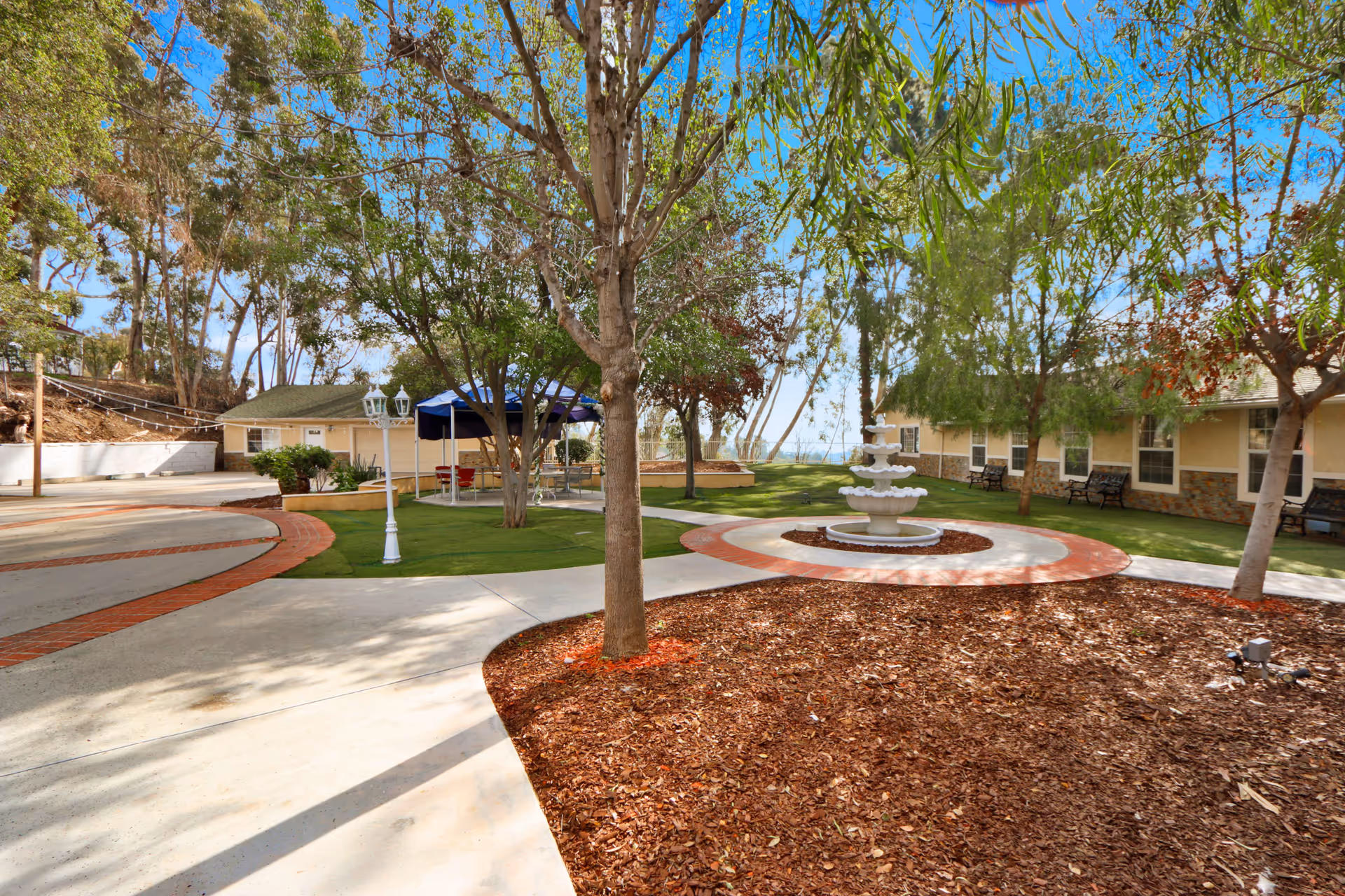 Courtyard with a central tiered fountain, walking paths, trees, benches, and a covered seating area.