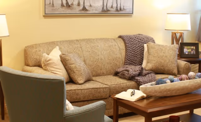 A cozy living room with a patterned beige sofa adorned with pillows and a knitted throw blanket. In front of the sofa is a wooden coffee table with an open book and a decorative bowl filled with colorful spheres. A blue upholstered chair is partially visible in the foreground. The room is softly lit by two table lamps on either side of the sofa, and a framed picture and a painting of trees hang on the wall.