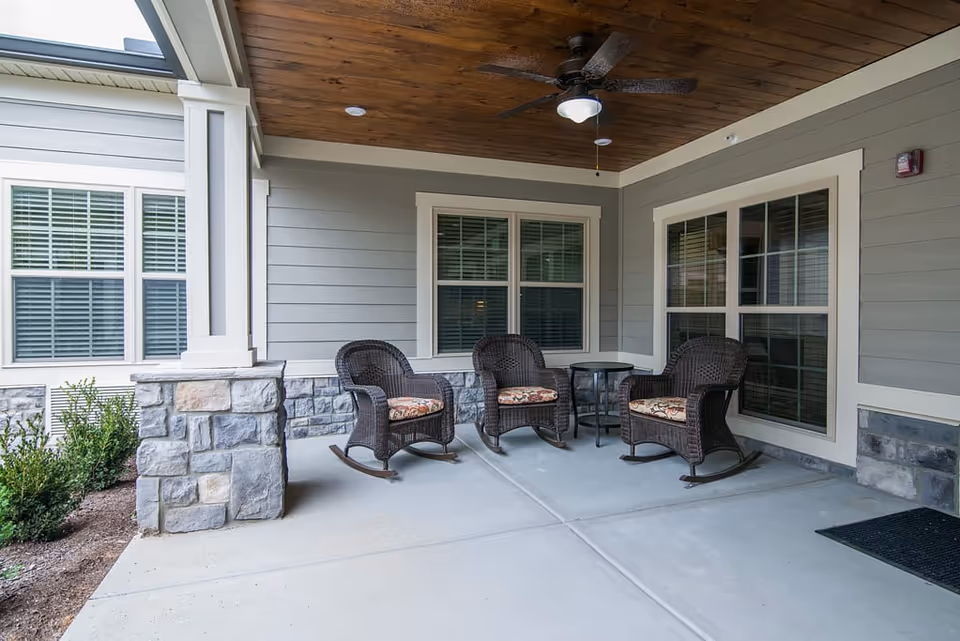 Covered outdoor patio area with three wicker rocking chairs with patterned cushions arranged around a small round table. The patio has a wooden ceiling with a ceiling fan and light, stone pillars, and large windows with white trim.