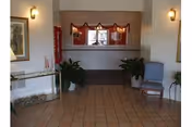 Interior view of a hallway or lobby area with tiled floor, two wall-mounted lights, a glass table with decorative items on the left, two potted plants flanking a window with red garland decorations, and a blue upholstered chair on the right.