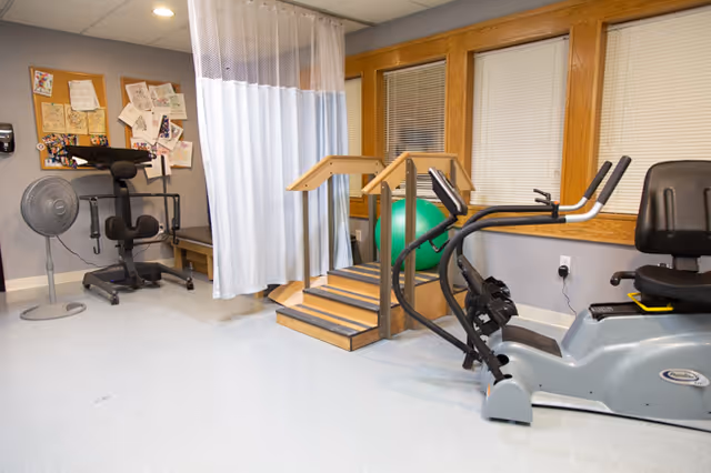 A physical therapy or rehabilitation room with exercise equipment including a recumbent bike, a set of wooden stairs with handrails, a green exercise ball, a standing fan, and a therapy chair. There is a white privacy curtain and a bulletin board with papers pinned on the wall.