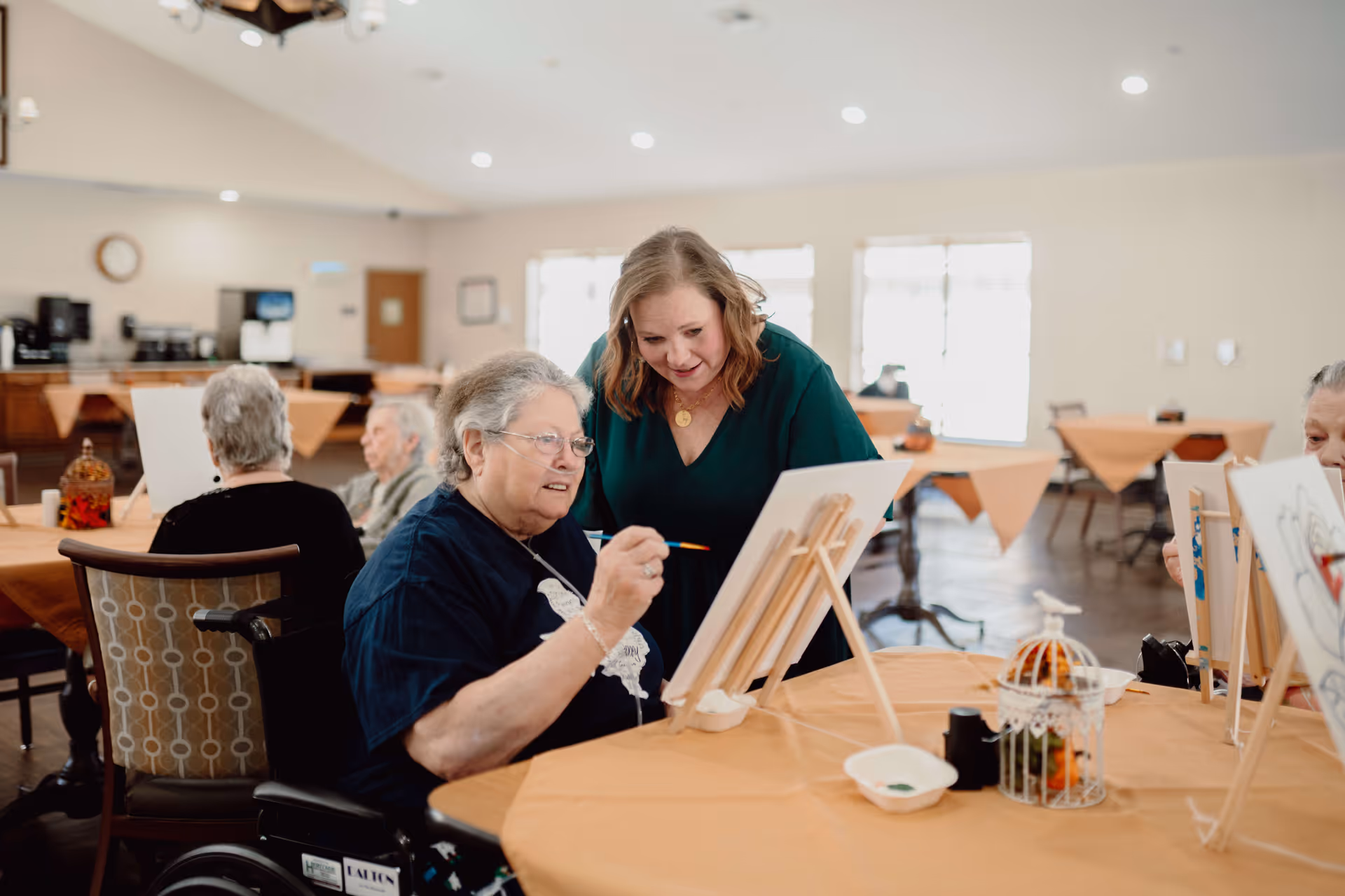 An elderly woman in a wheelchair painting on a canvas with the assistance of a younger woman in a green top in a well-lit room with tables covered in orange tablecloths. Other elderly individuals are also seated at tables, engaged in similar activities.