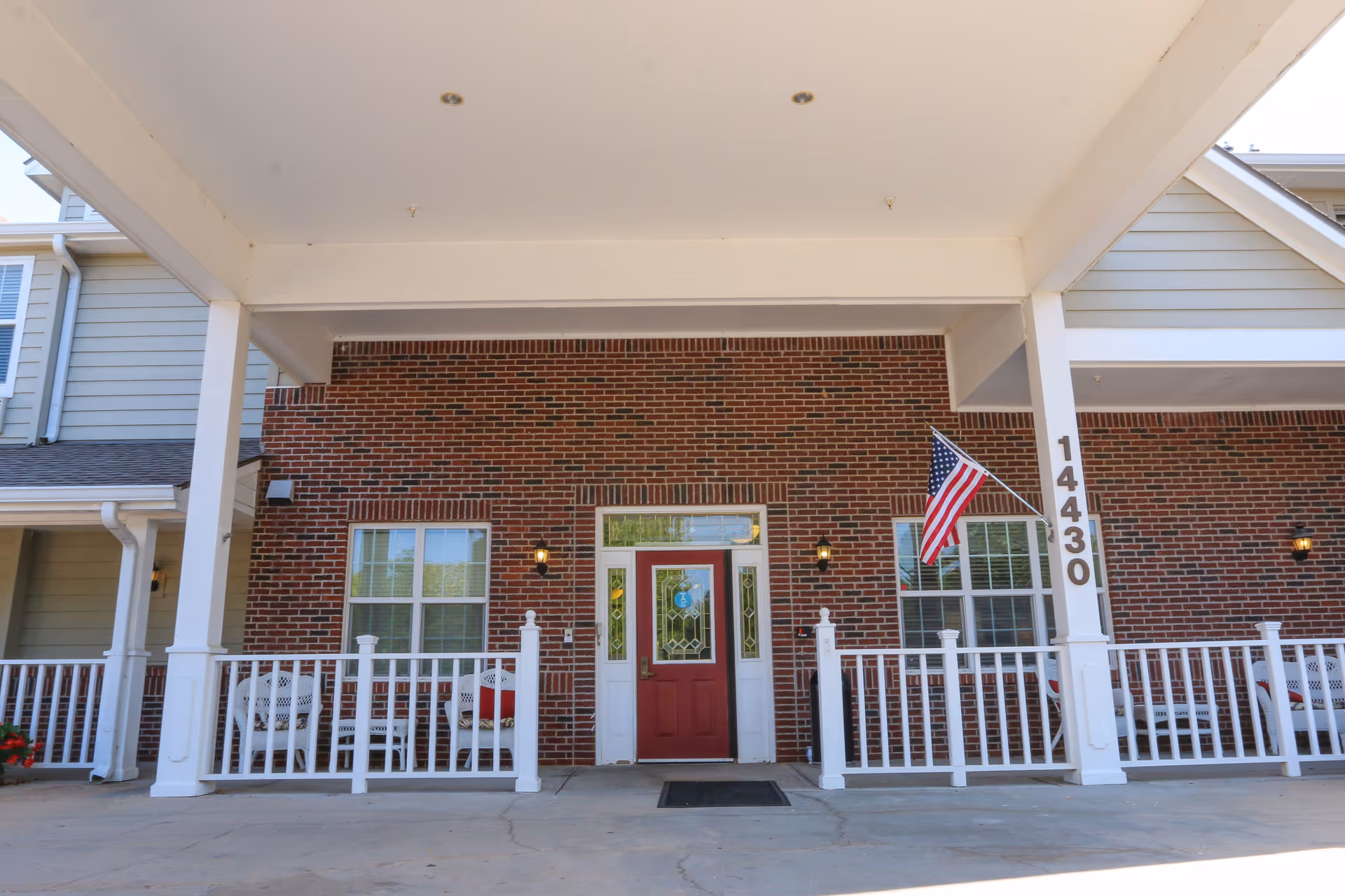 Front entrance of a brick building with a red door, white railing, and an American flag mounted on the right side. The building number 14430 is displayed vertically on a white pillar. There are white benches on the porch area.