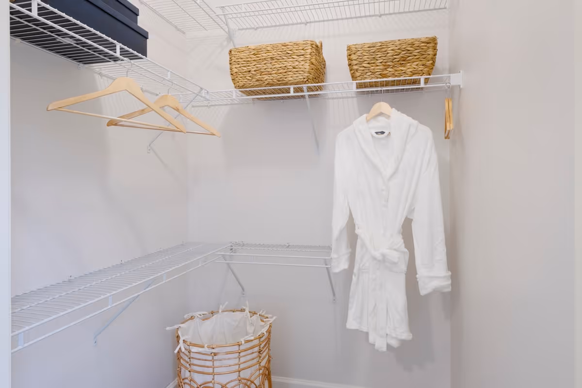 A small walk-in closet with white wire shelving. There are two wicker baskets on the top shelf, three wooden hangers on the left side, and a white bathrobe hanging on a hanger on the right side. A woven laundry basket with a white liner is on the floor.
