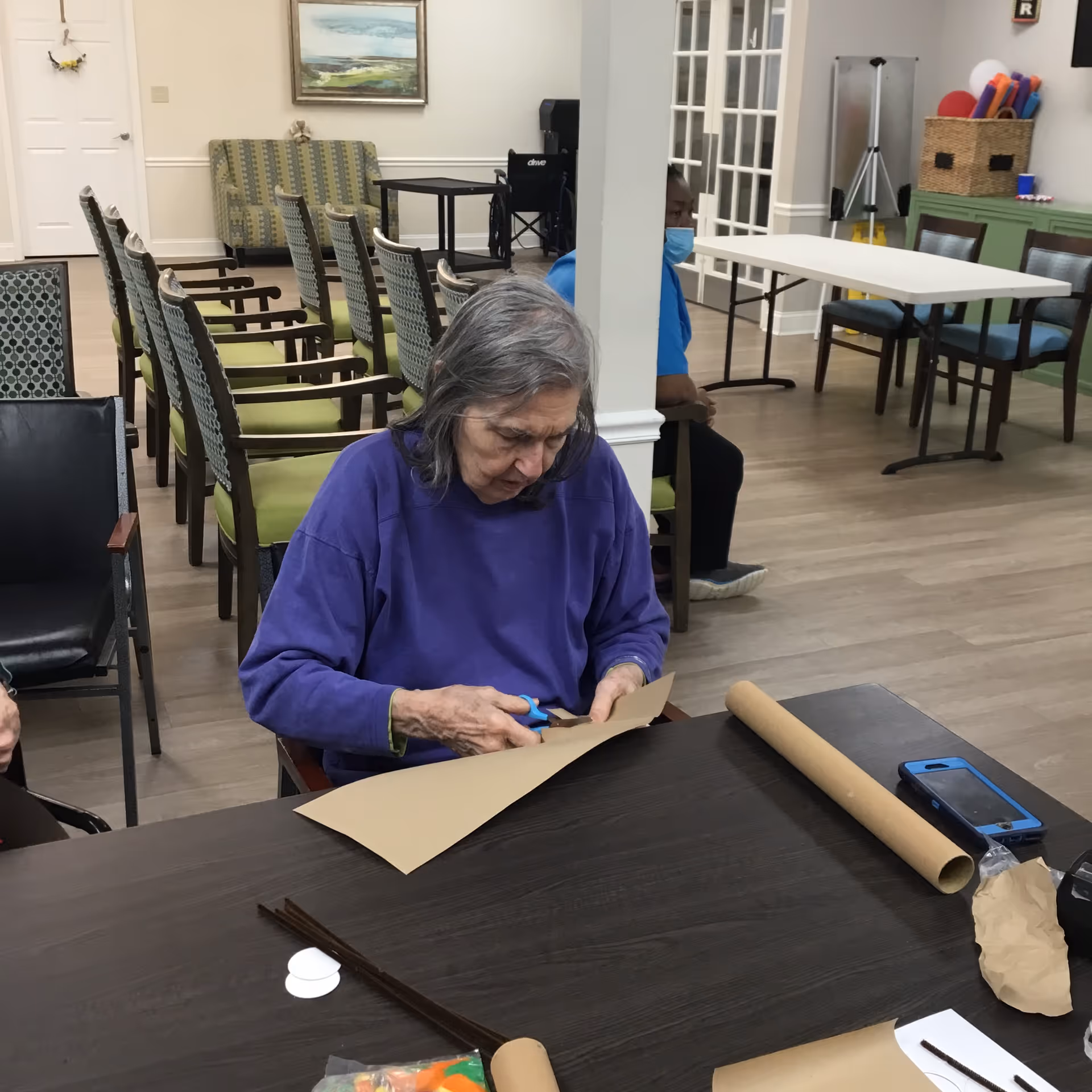 An elderly woman wearing a purple sweater is sitting at a table cutting brown paper with scissors in a room with multiple chairs and tables. Another person wearing a blue shirt and a face mask is seated in the background. The room has light-colored walls, a painting, and various items including a wheelchair and storage baskets.