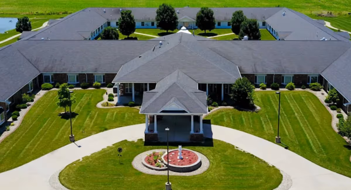 Aerial view of a single-story senior living facility with a large U-shaped building surrounding a central courtyard. The building has a gray roof and brick exterior with multiple windows. The courtyard features well-maintained green lawns, a circular driveway with a covered entrance, and a small landscaped area with a statue or fountain in the center.