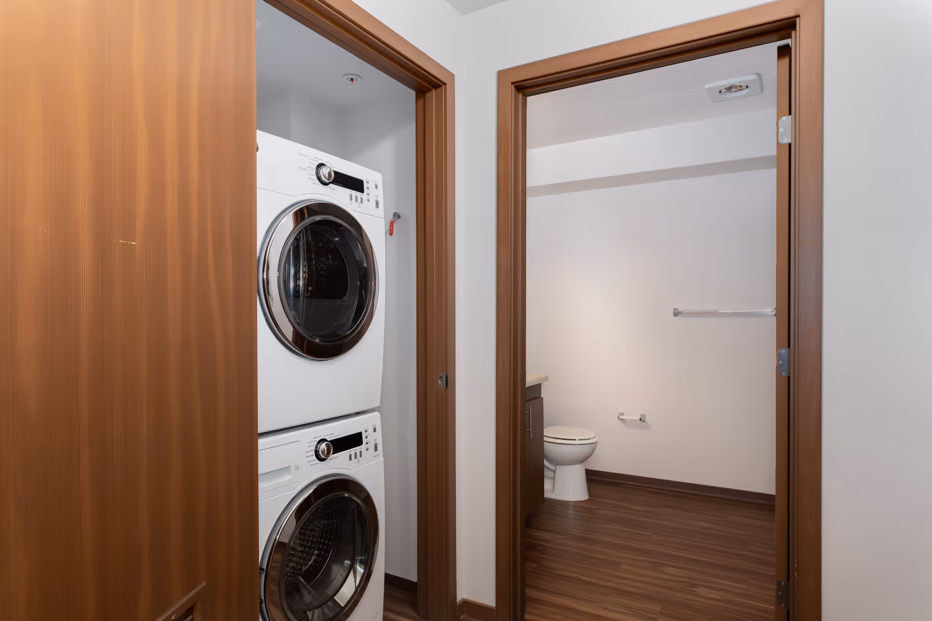 View of a laundry area with a stacked washer and dryer on the left and an adjacent bathroom with a toilet and a towel rack on the right, both rooms framed by wooden doorways and featuring wood flooring.