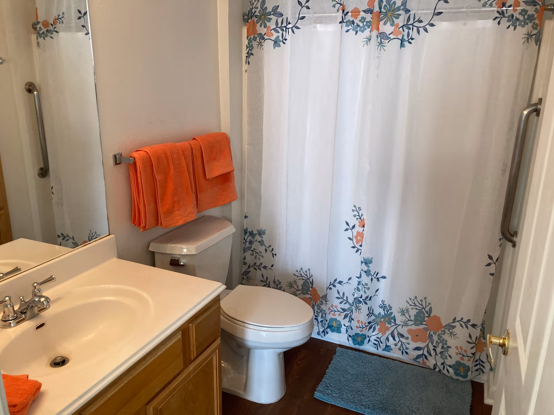 A bathroom with a white sink and wooden cabinet, a toilet with orange towels hanging above it, a shower with a white curtain decorated with blue and orange floral patterns, and a blue bath mat on the wooden floor.
