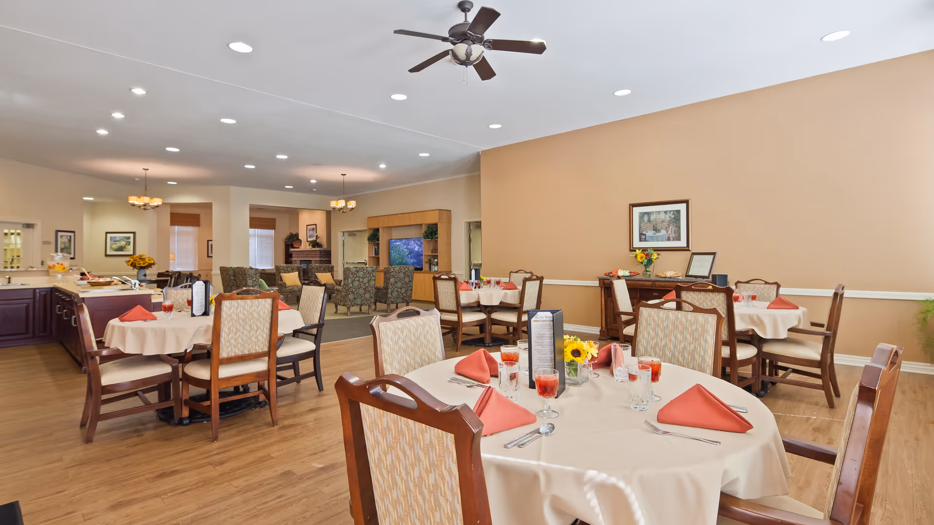 A spacious dining room with round tables covered in white tablecloths, each set with red folded napkins, glasses, and silverware. The room has wooden chairs with cushioned seats, hardwood floors, beige walls, and ceiling fans. In the background, there is a living area with patterned armchairs and a large TV mounted on a wooden entertainment center.