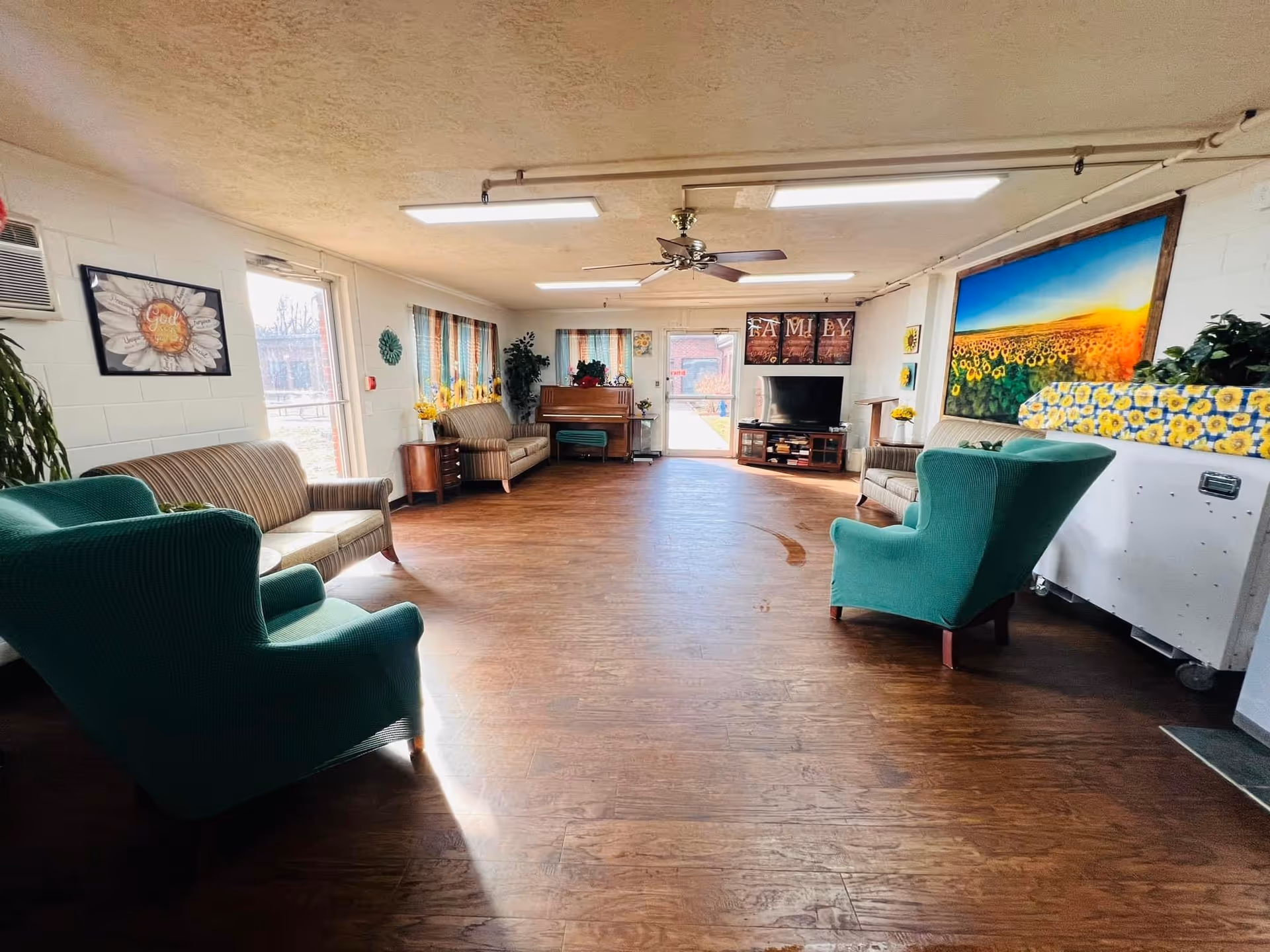 Bright communal living room with green armchairs and sofas arranged around a TV and piano, a large sunflower mural on the wall, and wood flooring.