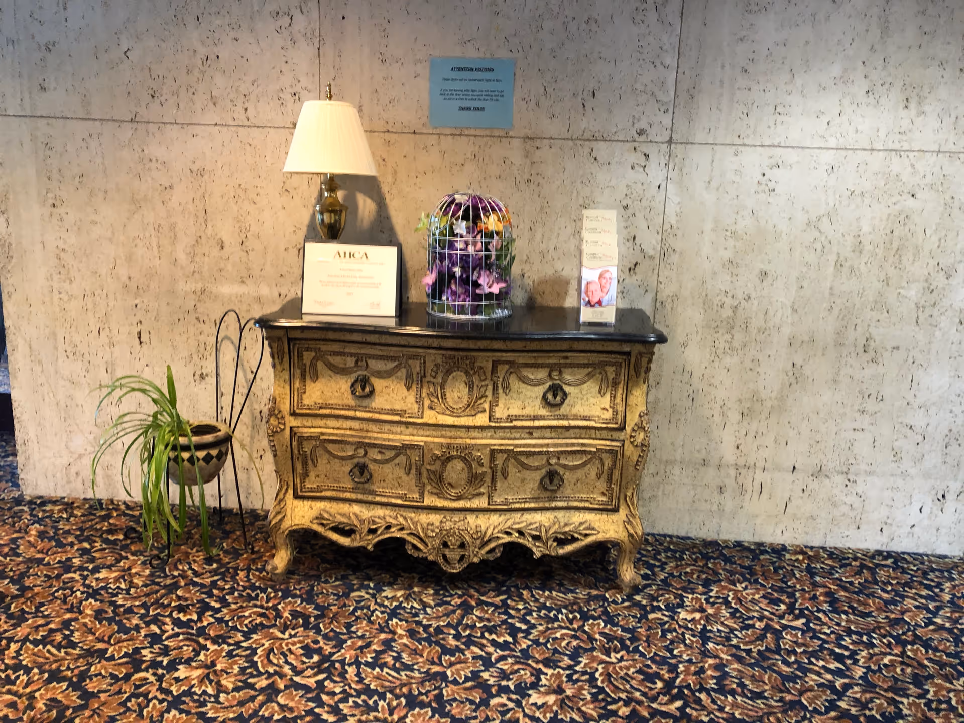 An ornate wooden dresser with two drawers stands against a textured wall. On top of the dresser is a table lamp with a white shade, a framed certificate, a decorative birdcage with purple flowers inside, and a small brochure holder with a pamphlet. To the left of the dresser is a potted plant on a metal stand. The floor is covered with a patterned carpet featuring autumn leaf designs.