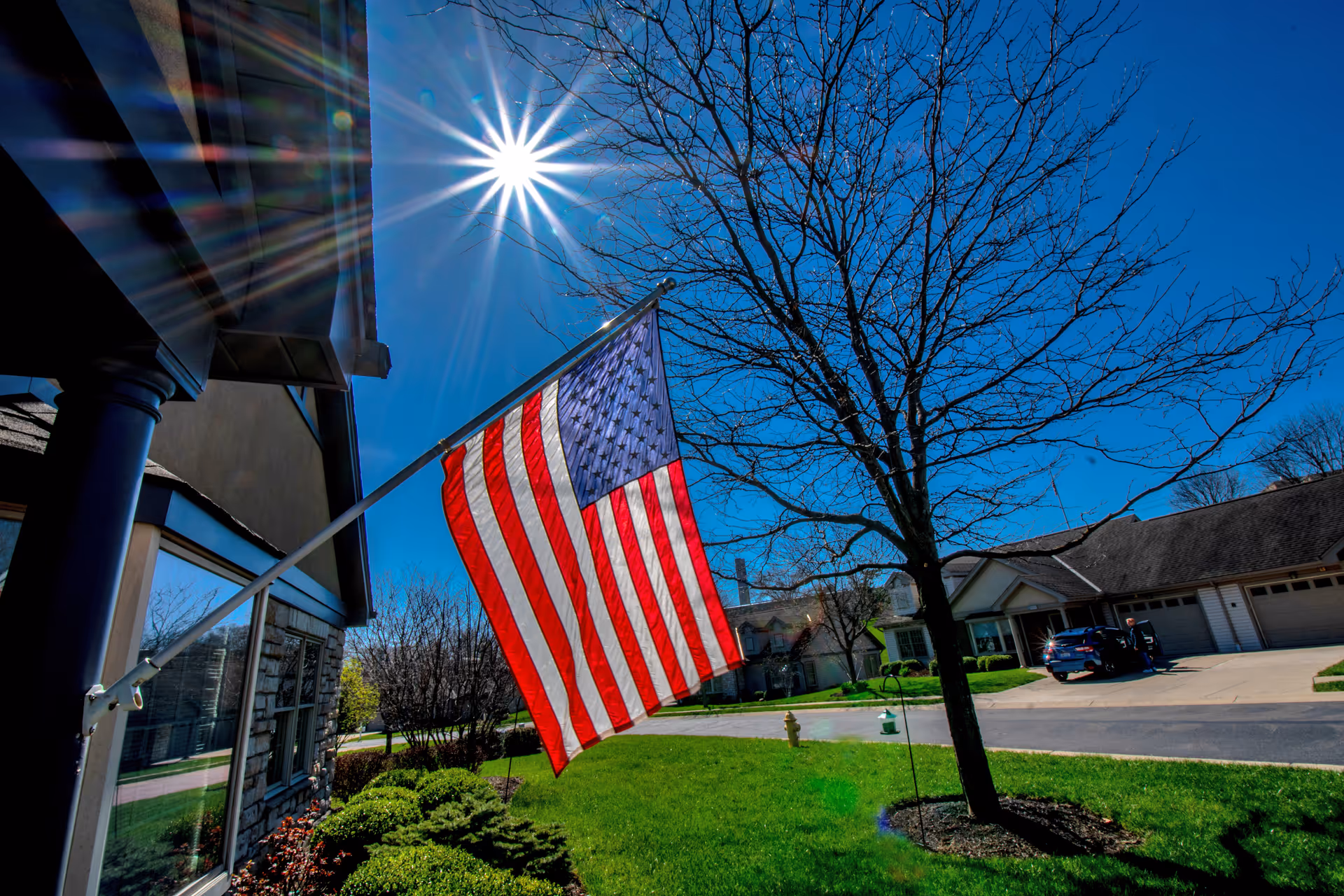 A bright sunny day with a clear blue sky, an American flag mounted on the side of a building waving in the breeze. In the foreground, there is a green lawn with a tree without leaves and some bushes. Across the street, there are residential-style buildings with garages and parked cars.