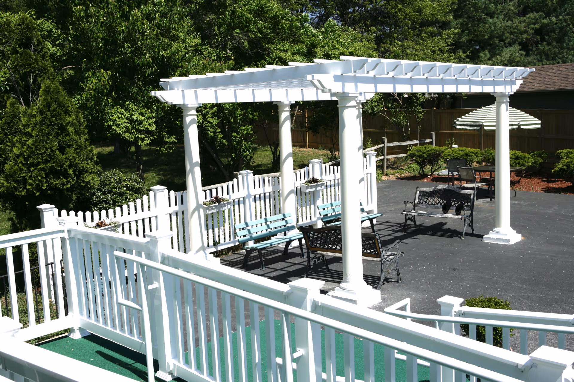 Sunny outdoor courtyard with a white pergola over benches and tables, white railings and picket fencing, and surrounding greenery.