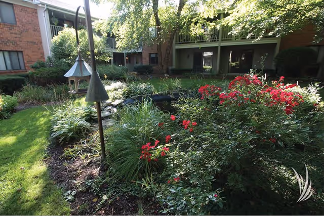 Landscaped courtyard with a small pond, red flowering bushes, a bird feeder, and brick apartment balconies in the background.