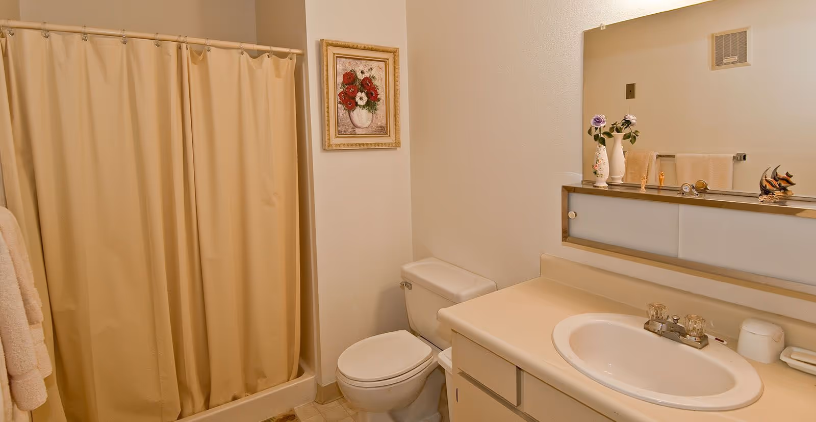A bathroom with a beige shower curtain, a white toilet, a countertop with a sink, and a large mirror above the sink. There are decorative items on the countertop including vases with flowers and small figurines. A framed floral painting hangs on the wall above the toilet.