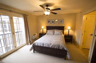 A cozy bedroom with a double bed centered against the wall, flanked by two nightstands each with a lamp. Above the bed are four framed pictures. To the left, there are large glass doors with curtains allowing natural light to enter. A ceiling fan with a light fixture is mounted above the bed. The room has beige walls and carpeted floor.
