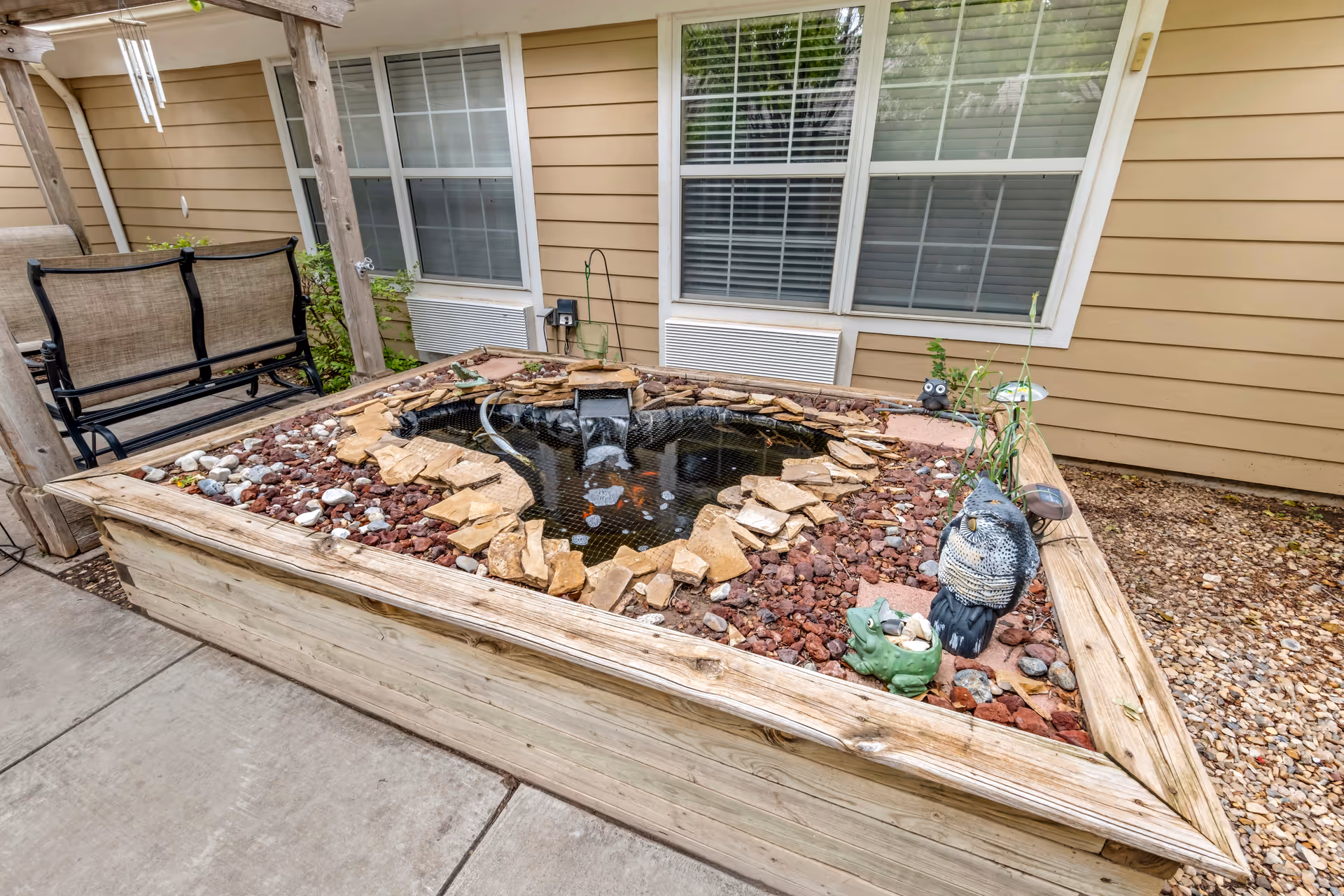 Raised wooden planter with a small decorative pond surrounded by rocks and garden ornaments in a courtyard outside a beige-sided building, with a bench swing and windows in the background.
