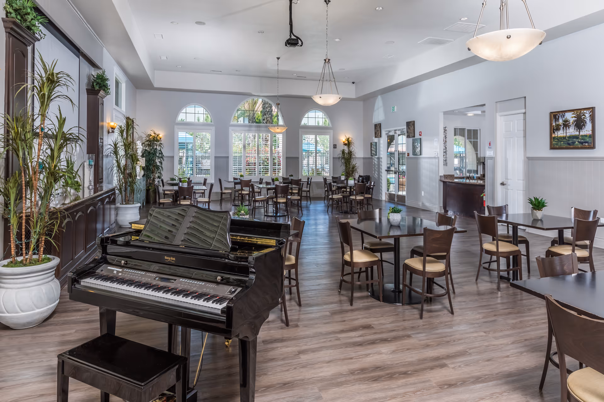 A spacious dining room with multiple tables and chairs arranged neatly. A black grand piano with a matching bench is positioned in the foreground. The room features large arched windows with white shutters allowing natural light to fill the space. There are several potted plants and wall decorations, with light fixtures hanging from the ceiling.