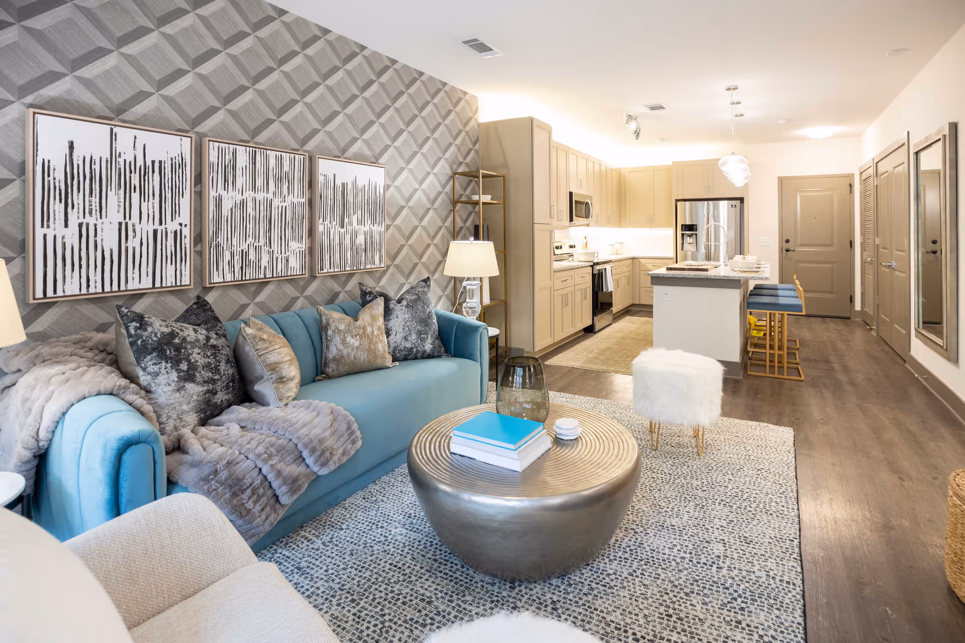 A modern open-concept living room and kitchen area featuring a light blue sofa with multiple decorative pillows and a gray throw blanket. The living room has a round metallic coffee table with books and decor on top, a patterned rug, and a textured accent wall with three abstract black and white framed artworks. The kitchen has beige cabinets, stainless steel appliances, an island with a granite countertop, and three blue bar stools. The flooring is wood, and the space is well-lit with ceiling lights.
