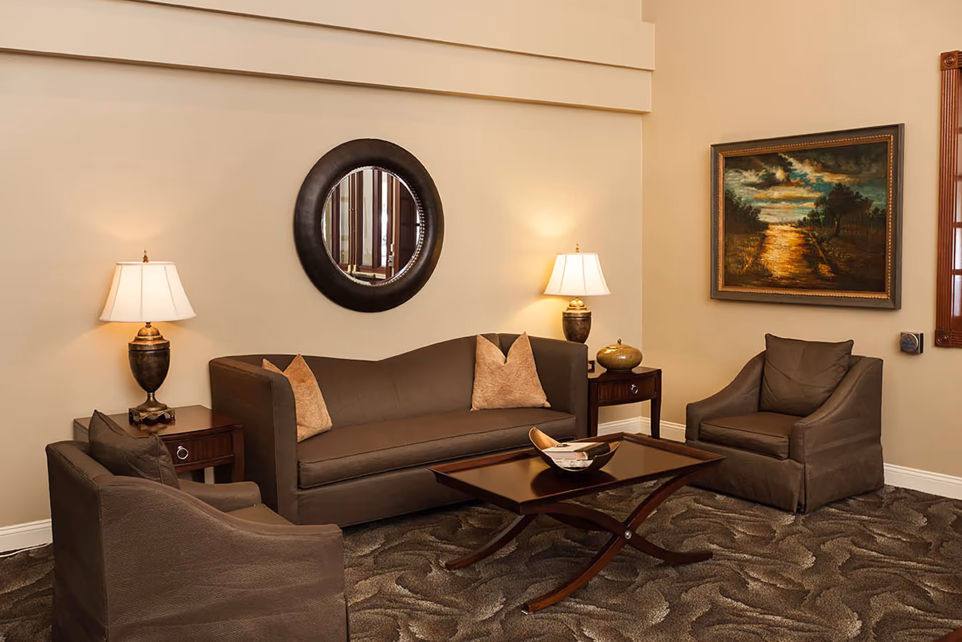 A cozy living room with a brown sofa and two matching armchairs arranged around a wooden coffee table. Two side tables with lamps flank the sofa. A round mirror hangs on the wall above the sofa, and a framed painting of a sunset over a river is on the adjacent wall. The room has beige walls and a patterned carpet.