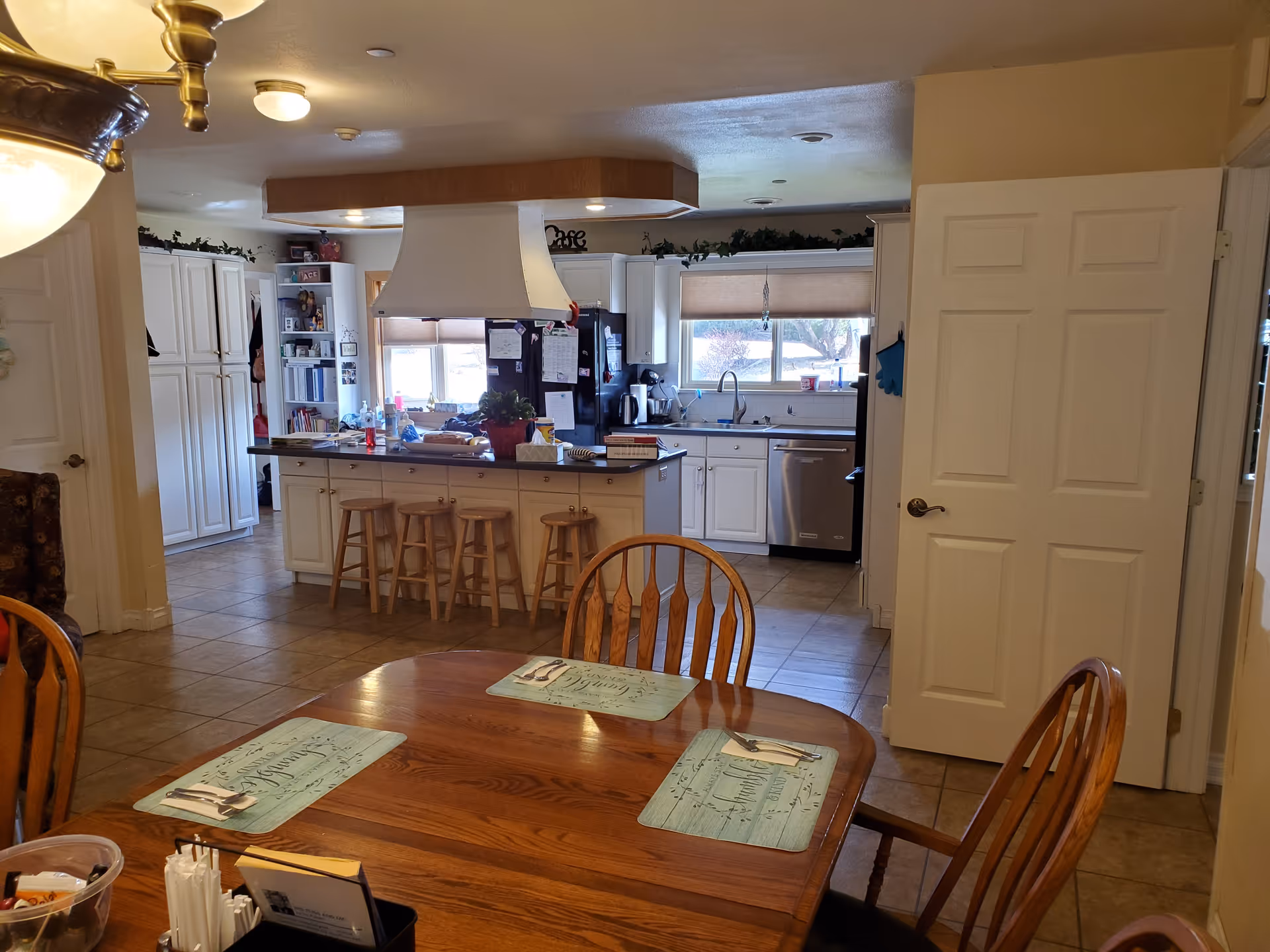 Wood dining table with placemats in the foreground and a spacious kitchen with island, stools, white cabinets, and a large range hood in the background.