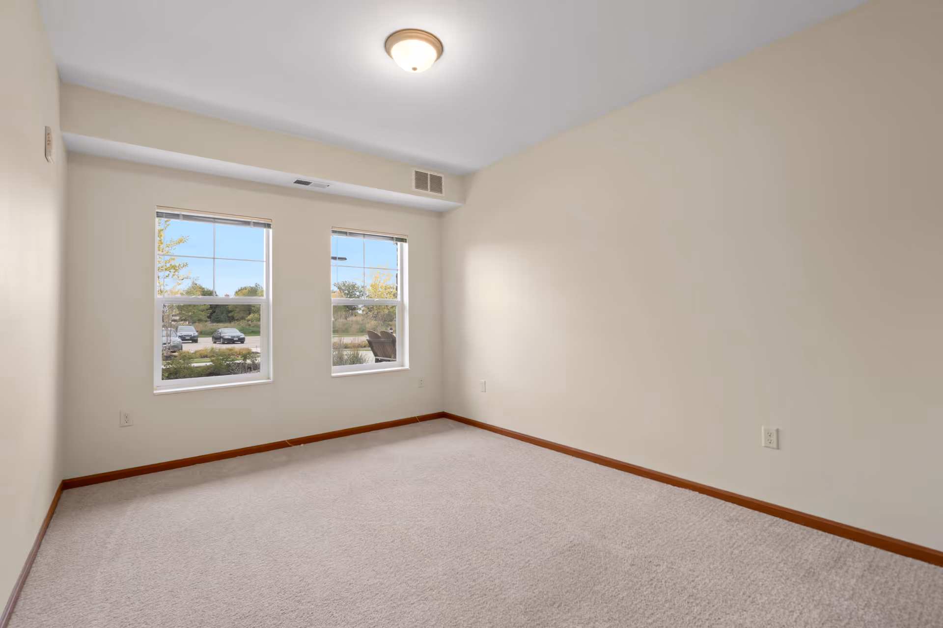Empty room with beige walls and carpeted floor, two windows showing a view of parked cars and greenery outside, ceiling light fixture, and wooden baseboards.