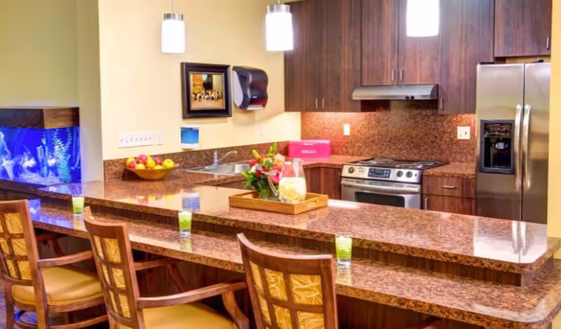 Modern memory care kitchen with a long granite breakfast bar, four wooden bar stools, stainless steel appliances, pendant lights, and a blue-lit fish tank.