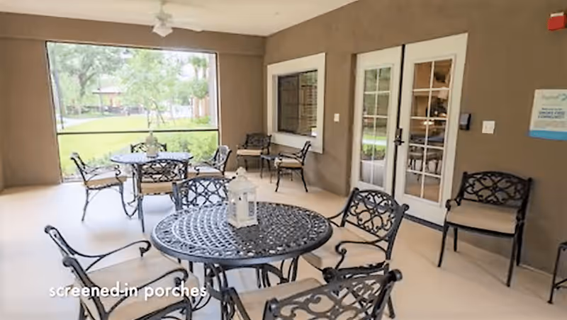Screened-in porch area with multiple black metal tables and chairs with intricate designs. The porch overlooks a green outdoor space with trees and a gazebo. There are double glass doors leading inside the building and additional seating along the wall.