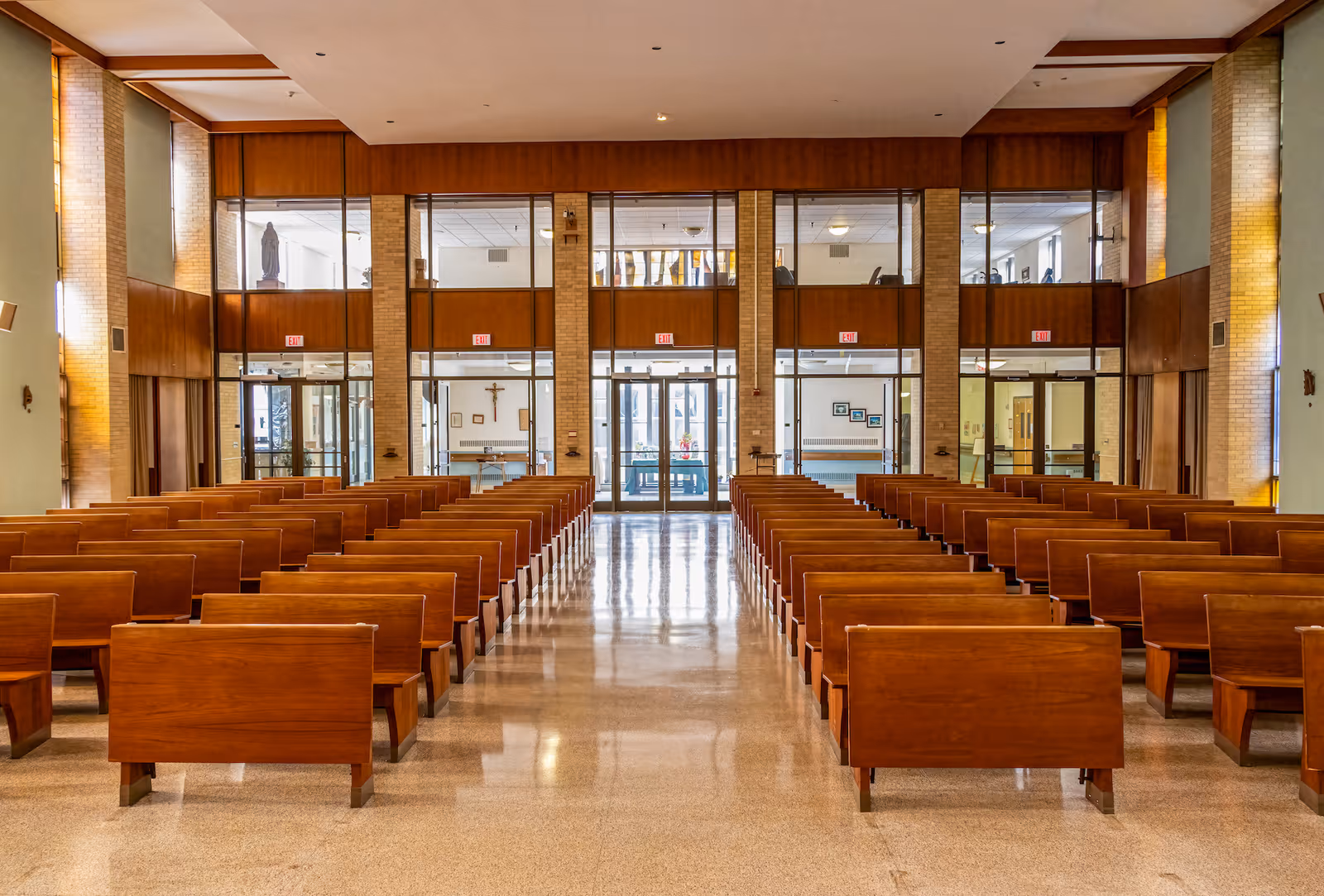 Interior view of a chapel or worship area with rows of wooden pews facing a glass wall with multiple doors and windows. The space is well-lit with natural light coming through the windows, and religious symbols such as a crucifix and a statue are visible in the background.