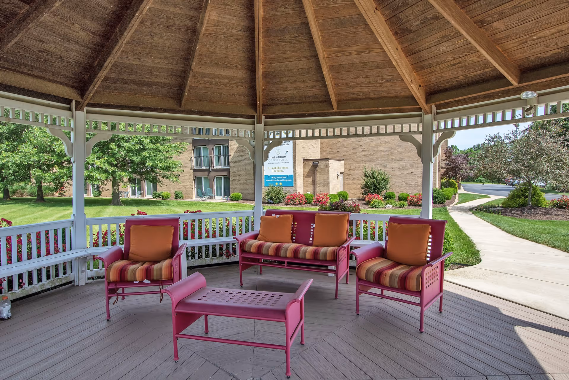 Outdoor gazebo with a wooden roof and white railing, furnished with red metal chairs and a loveseat with striped cushions and orange pillows. Surrounding the gazebo is a well-maintained garden with green grass, bushes, and trees, and a paved walkway leading away from the structure. A building and a sign for The Atrium of Belleville are visible in the background.